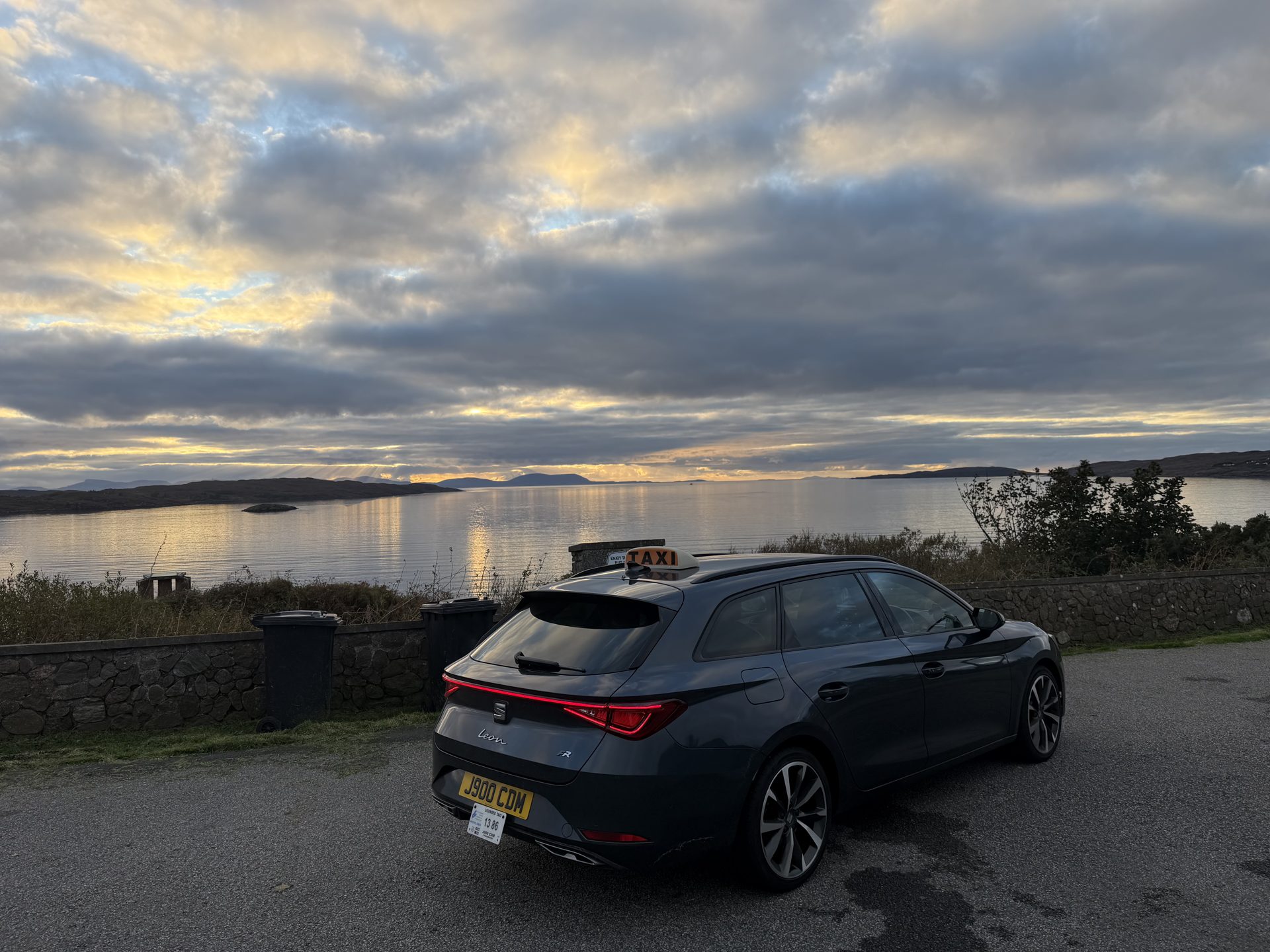 A West Coast Taxis car parked above a calm sea loch at sunset